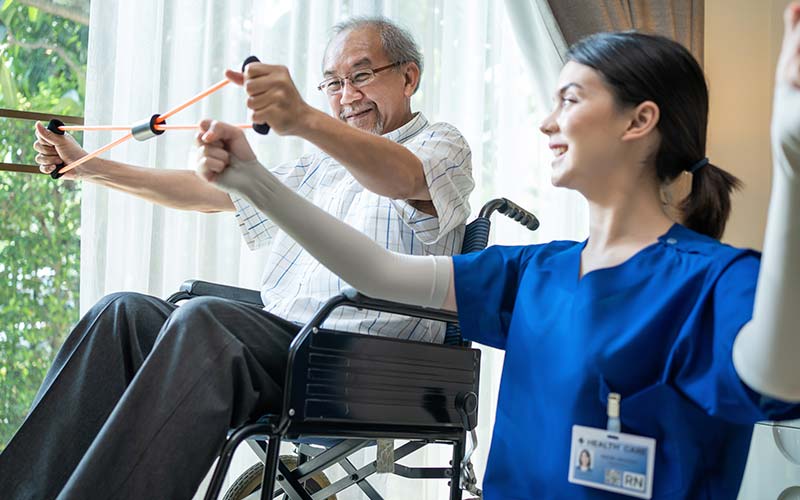 an senior man is in a wheelchair using resistance bands with the help of a therapeutic nurse