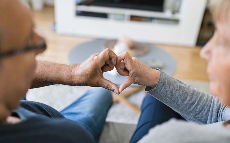 two seniors sitting on a couch in living room with both hands coming together for a heart shape