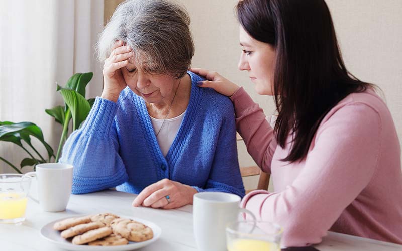 daughter consoling her mother who at a table with coffee and cookies as a way to exhibit an example of anosognosia