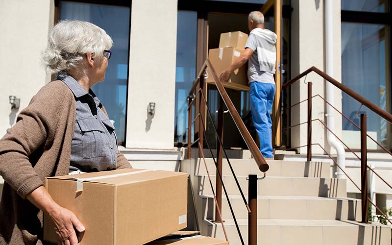 a senior couple carries moving boxes into their smaller home they downsized to