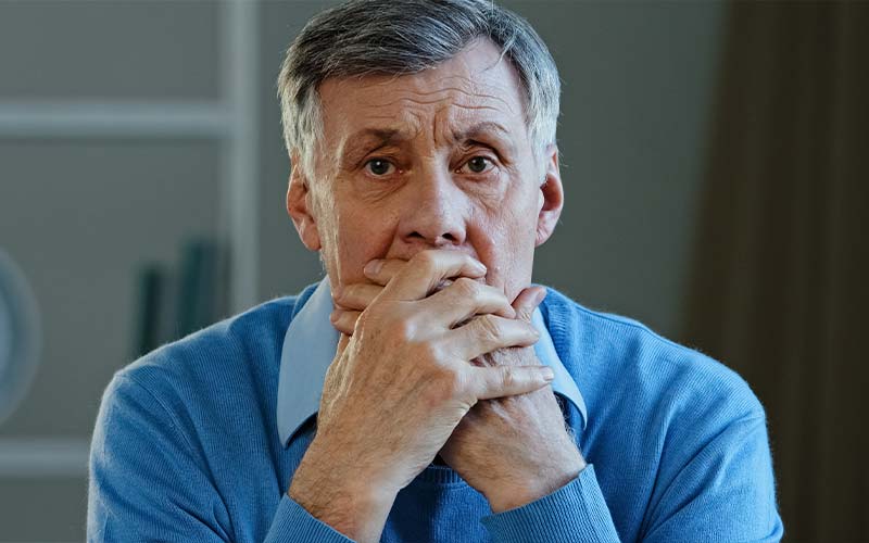 a worried senior man stares at the camera with both hands covering his mouth to highlight the toll elder abuse can take
