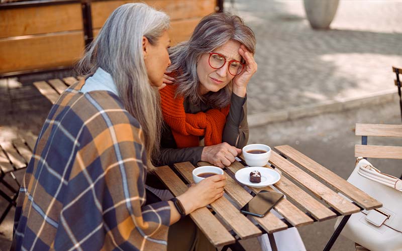 a senior woman consoles another senior woman at a park table