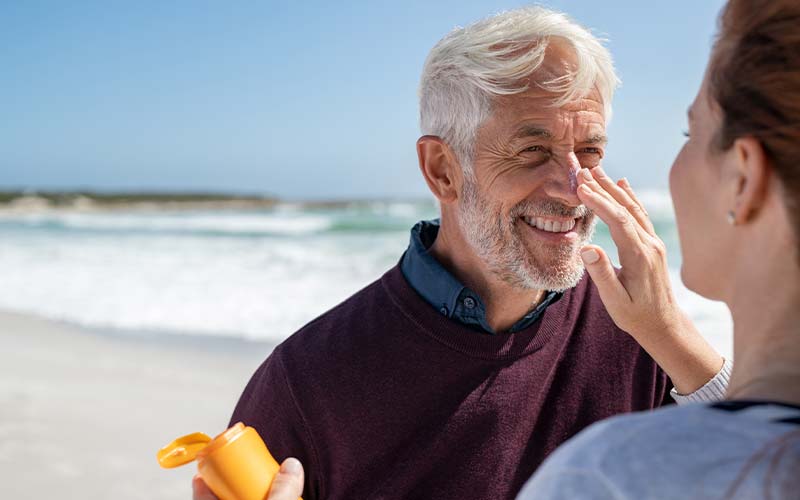 a young woman puts sunscreen on her senior father at the beach to protect from uv rays