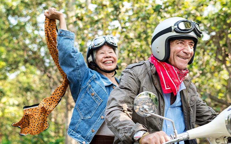 a senior couple on a scooter with helmets and big smiles, covered and protected from uv rays